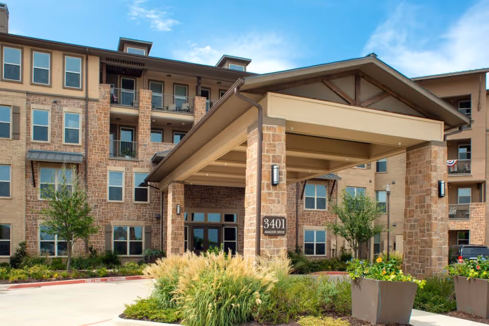 Front exterior view of a multi-story senior living facility building with a covered entrance supported by stone pillars, surrounded by landscaped greenery and plants under a partly cloudy sky.