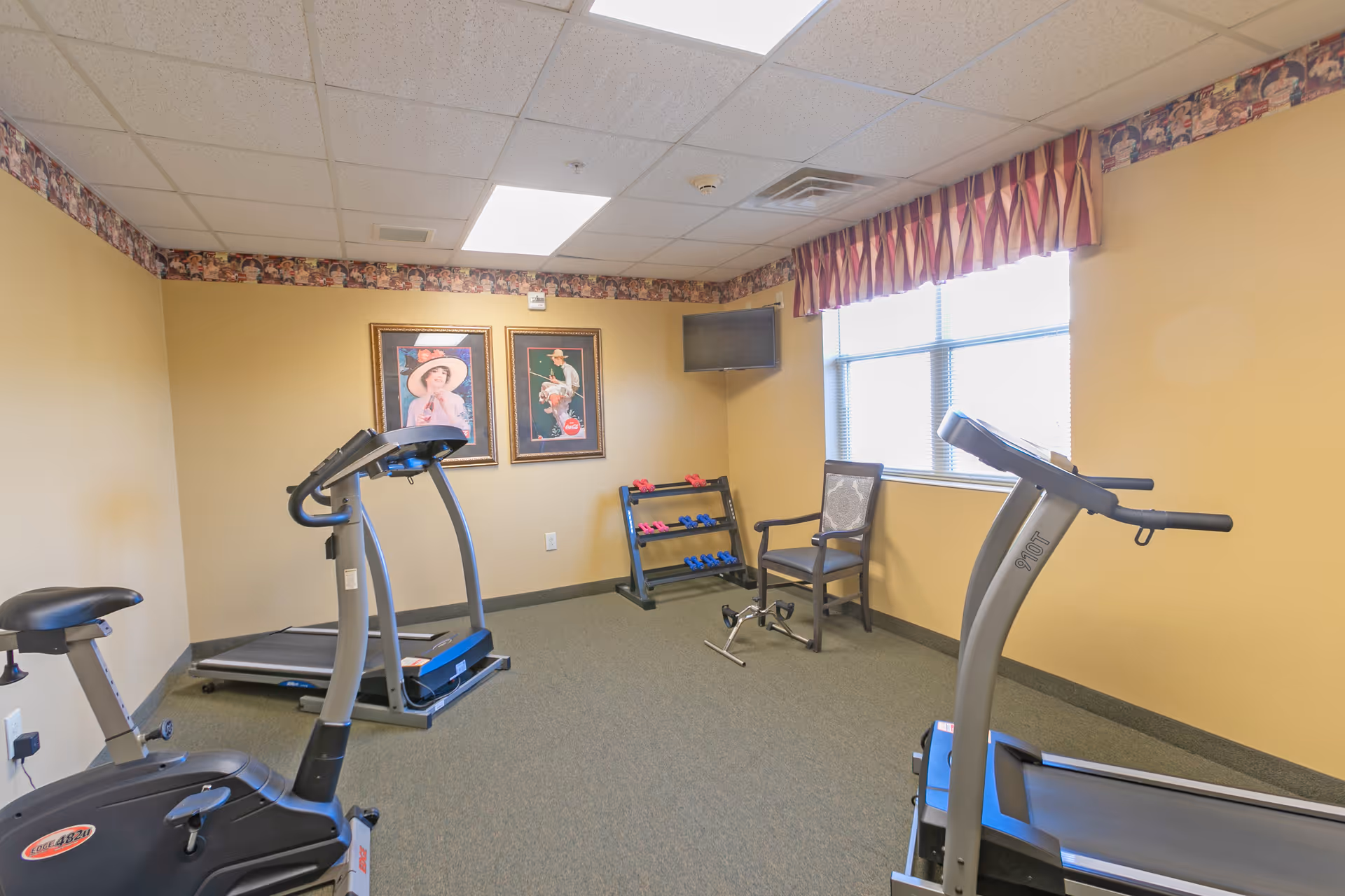 A small exercise room with two treadmills, a stationary bike, a chair, a rack of dumbbells, two framed pictures on the wall, and a wall-mounted TV. The room has beige walls, a window with a striped valance, and a carpeted floor.