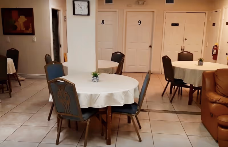 Interior view of a common area with round tables covered with white tablecloths, each table having a small potted plant. Several chairs are arranged around the tables. There are doors numbered 8 and 9 in the background, a wall clock, and a brown leather couch on the right side.