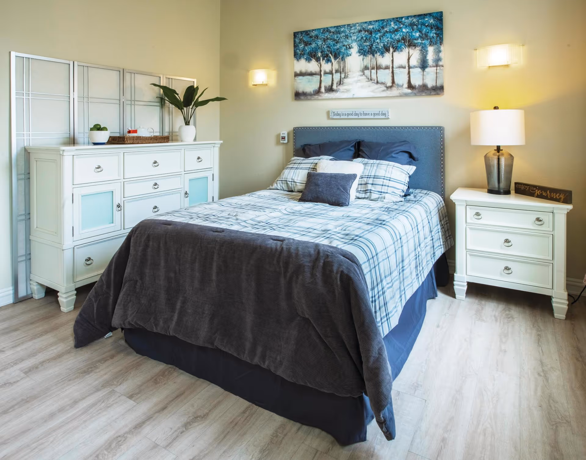 A neatly made bed with plaid bedding and multiple pillows in a senior living bedroom. The room features light-colored wooden flooring, a white dresser with decorative items including a plant and a bowl of green apples, and a matching white nightstand with a lamp and a small sign that reads 'enjoy the journey'. Above the bed is a painting of a tree-lined path and a small sign that says 'Today is a good day to have a good day'. The walls are painted a soft beige color.
