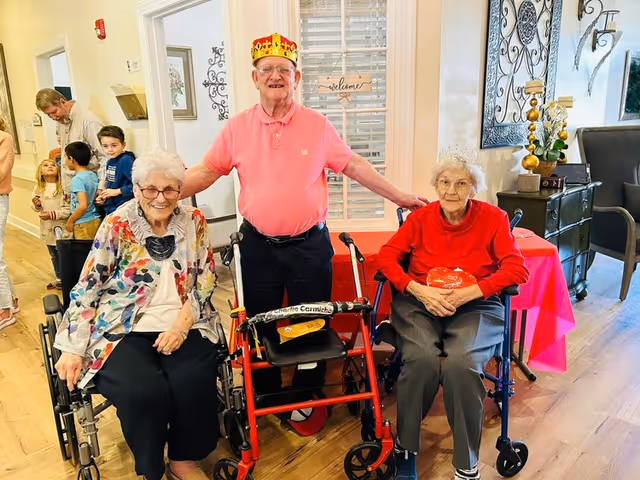 Three elderly individuals in a senior living facility common area. Two women are seated in wheelchairs, one wearing a colorful floral jacket and the other in a red sweater with a tiara. A man stands between them wearing a pink shirt and a red and gold crown, with his arms around the women. In the background, several children and adults are visible near a hallway. The room has wooden flooring, a decorative wall piece, and a table with a pink tablecloth.