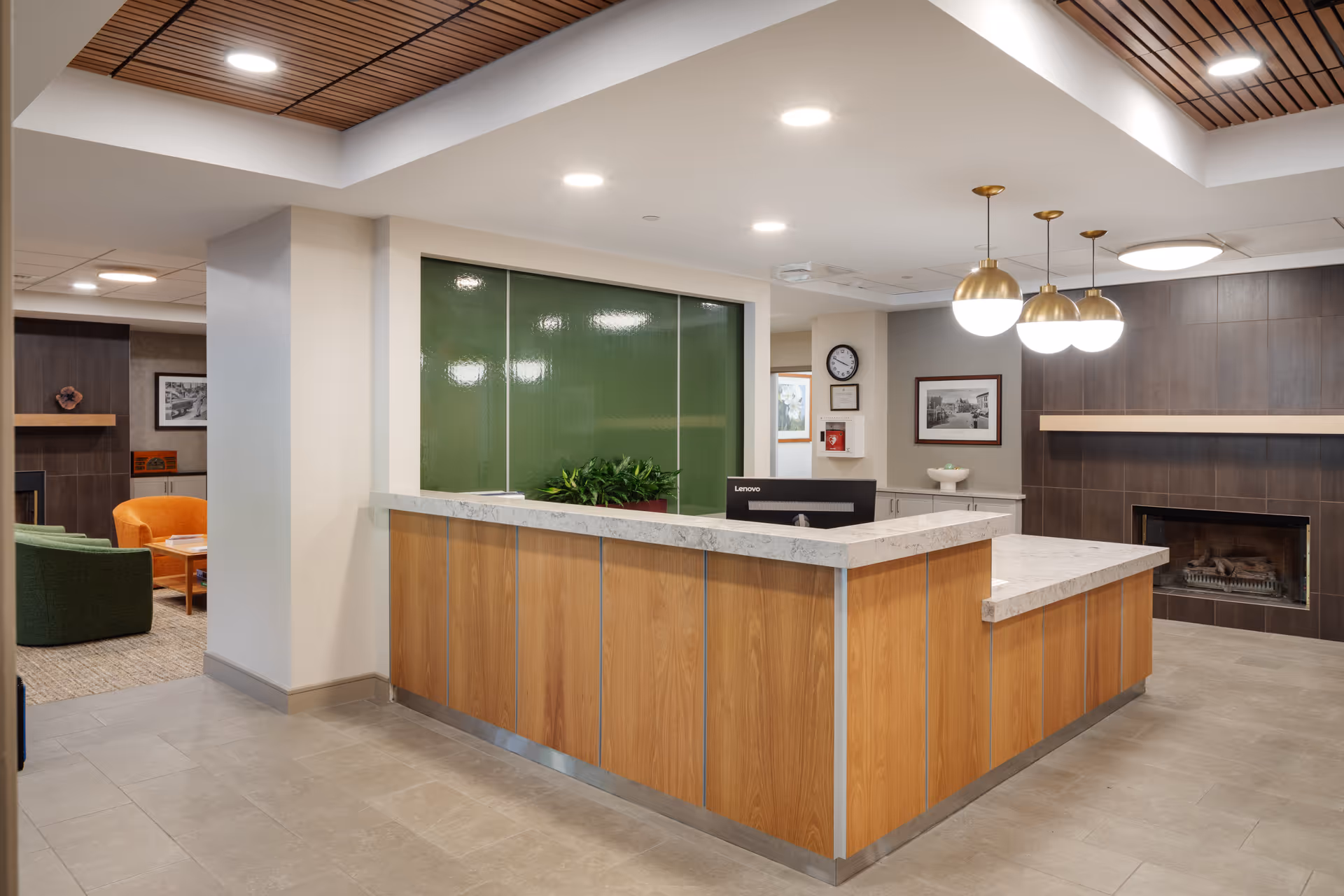 Reception area in a senior living facility with a wooden and marble front desk, a green glass panel behind the desk, and a computer monitor. The ceiling has wooden slats and recessed lighting. To the right, there is a fireplace with a dark tile surround and three hanging globe pendant lights. In the background, there is a seating area with green and orange chairs and framed pictures on the walls.