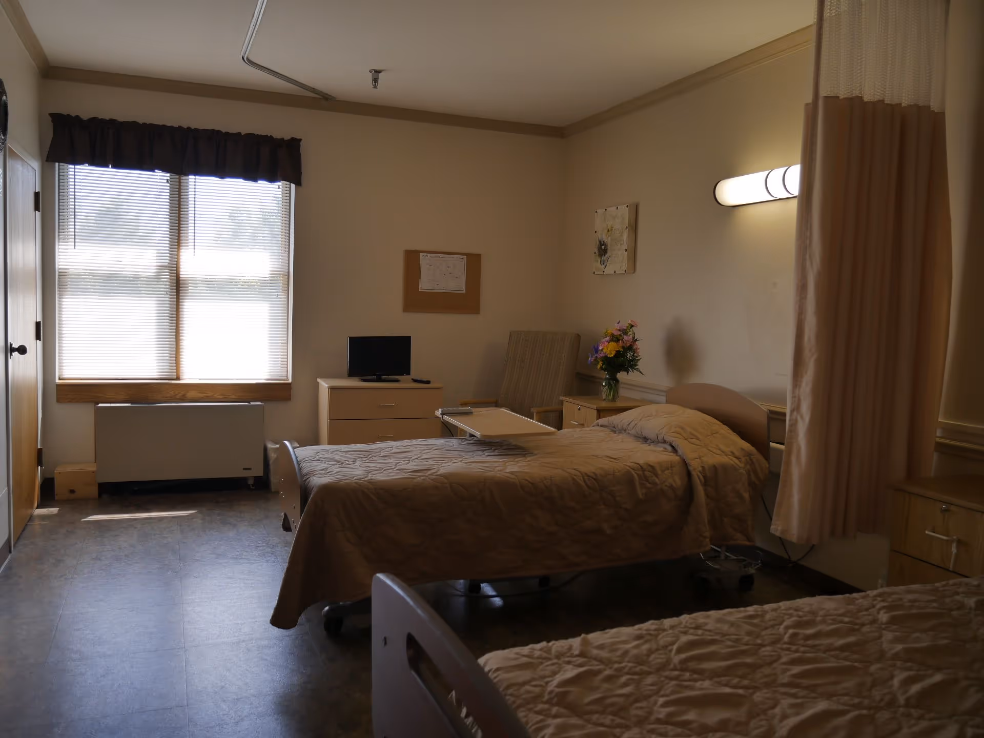 A healthcare facility room with two hospital beds covered in beige quilts. There is a window with blinds and a dark valance, a small TV on a dresser, a chair, a side table with a vase of flowers, and a privacy curtain partially drawn. The room has neutral-colored walls and flooring.