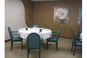A dining room with a round table covered with a white tablecloth and set with plates, cups, and utensils. Six teal upholstered chairs surround the table. The walls are brown with two framed floral paintings hanging on them.