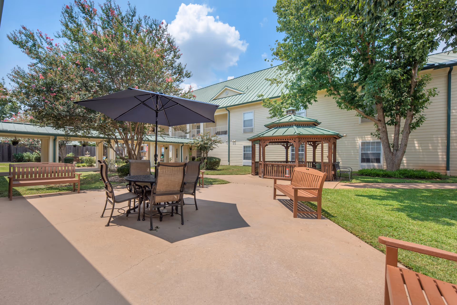 Outdoor patio area at Brookdale Beckett Meadows featuring a round table with four chairs under a large black umbrella, wooden benches, a gazebo with a green roof, trees, and a two-story building with a green metal roof in the background under a partly cloudy sky.