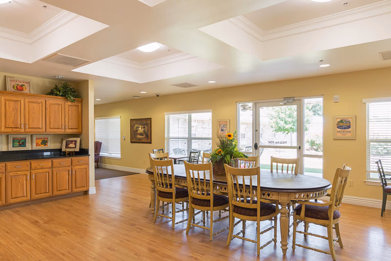 A bright dining room with a long wooden table surrounded by eight wooden chairs with cushioned seats. A floral centerpiece with sunflowers decorates the table. The room has light-colored walls, large windows with blinds, and a glass door labeled 'NOT AN EXIT' leading outside. Wooden cabinets with decorative items are on the left side, and the floor is light wood.