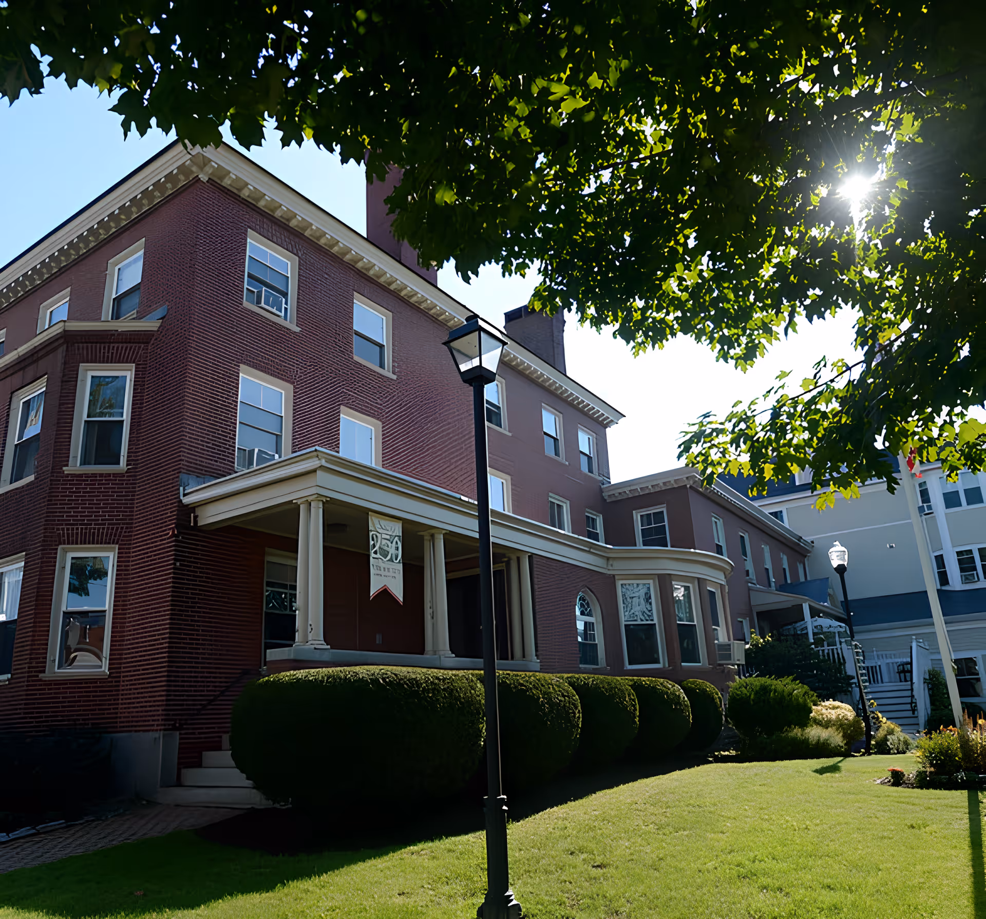Exterior view of a large brick building with multiple windows and a covered entrance supported by columns. The building is surrounded by neatly trimmed bushes and a well-maintained lawn. Sunlight filters through the green leaves of a tree overhead, casting shadows on the scene.