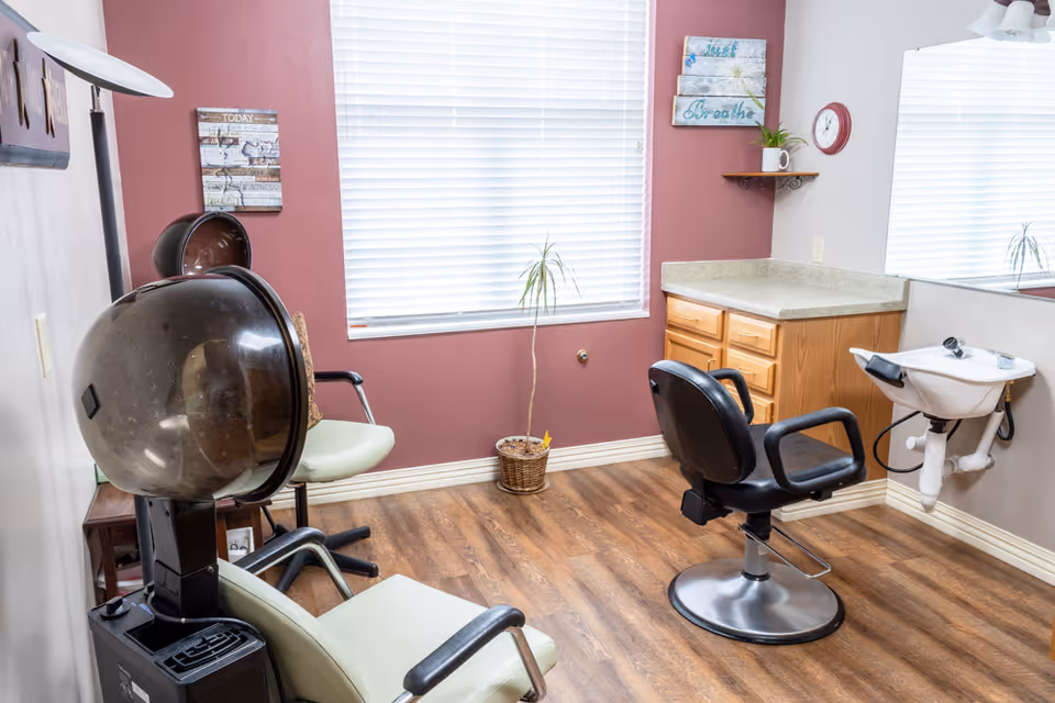 Interior of a small salon area with a hair dryer chair, a black salon chair in front of a sink, wooden cabinets, a window with blinds, and wall decorations including a clock and framed signs.