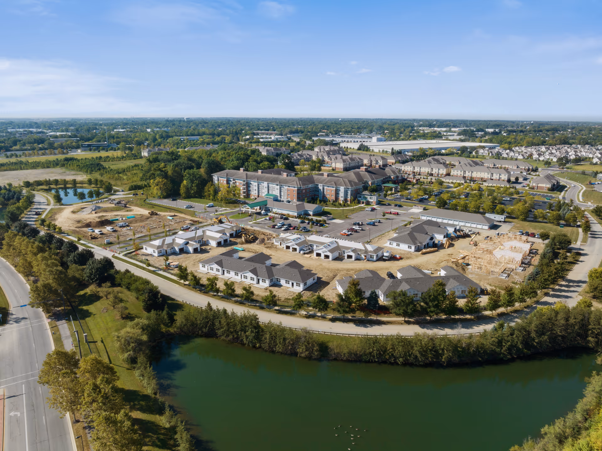 Aerial view of Verena at Hilliard senior living facility showing multiple buildings, parking lots, surrounding roads, greenery, and a pond in the foreground under a clear blue sky.