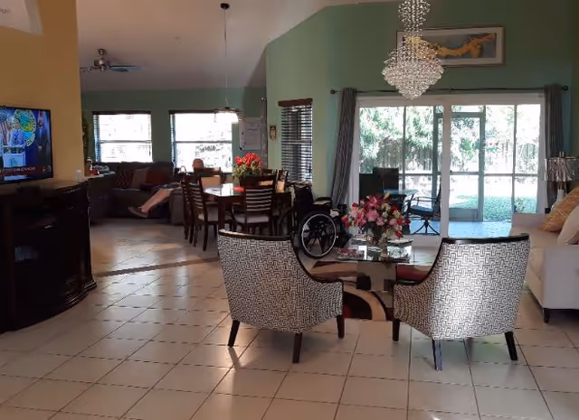 A spacious living and dining area with tiled floors. The foreground features two patterned armchairs around a glass coffee table with a flower arrangement. In the background, there is a dining table with chairs and a wheelchair nearby. Large sliding glass doors lead to an outdoor patio with greenery visible outside. A television is mounted on the left wall, and a chandelier hangs from the ceiling.