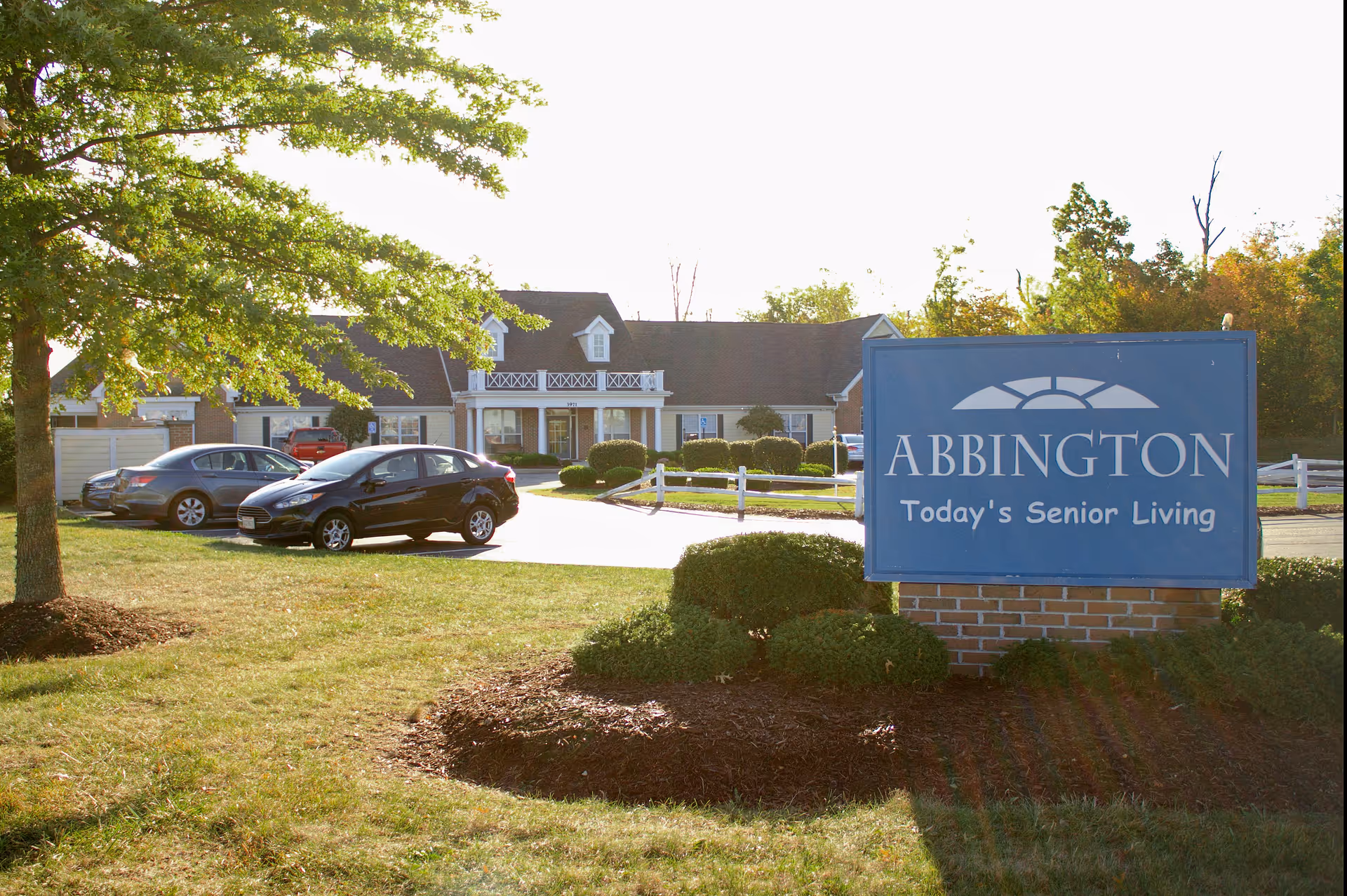 Exterior view of Abbington of Powell Assisted Living facility with a blue sign in the foreground that reads 'ABBINGTON Today's Senior Living'. The building is a single-story structure with a porch and white railings. Several cars are parked in the parking lot, and there are trees and bushes around the area.