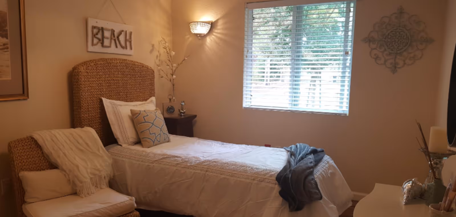 A cozy bedroom with a single bed featuring a woven headboard, white bedding, and decorative pillows. A soft throw blanket is draped over the foot of the bed. Next to the bed is a small wooden nightstand with a clock and decorative items. A window with white blinds lets in natural light. The walls are decorated with a 'BEACH' sign and a decorative metal wall piece. A cushioned wicker chair with a white throw blanket is positioned beside the bed.