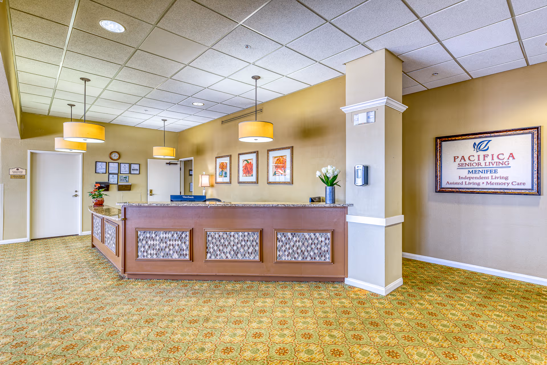Reception area of a senior living facility with a long wooden front desk, three hanging pendant lights, framed artwork on the wall, a clock, and a sign that reads 'Pacifica Senior Living Menifee Independent Living Assisted Living Memory Care'. The floor is covered with patterned carpet and there are flowers on the desk and a column.