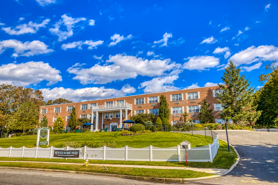 Front exterior view of a large brick assisted living facility named Amber Court with a well-maintained lawn, white picket fence, and a clear blue sky with scattered clouds.