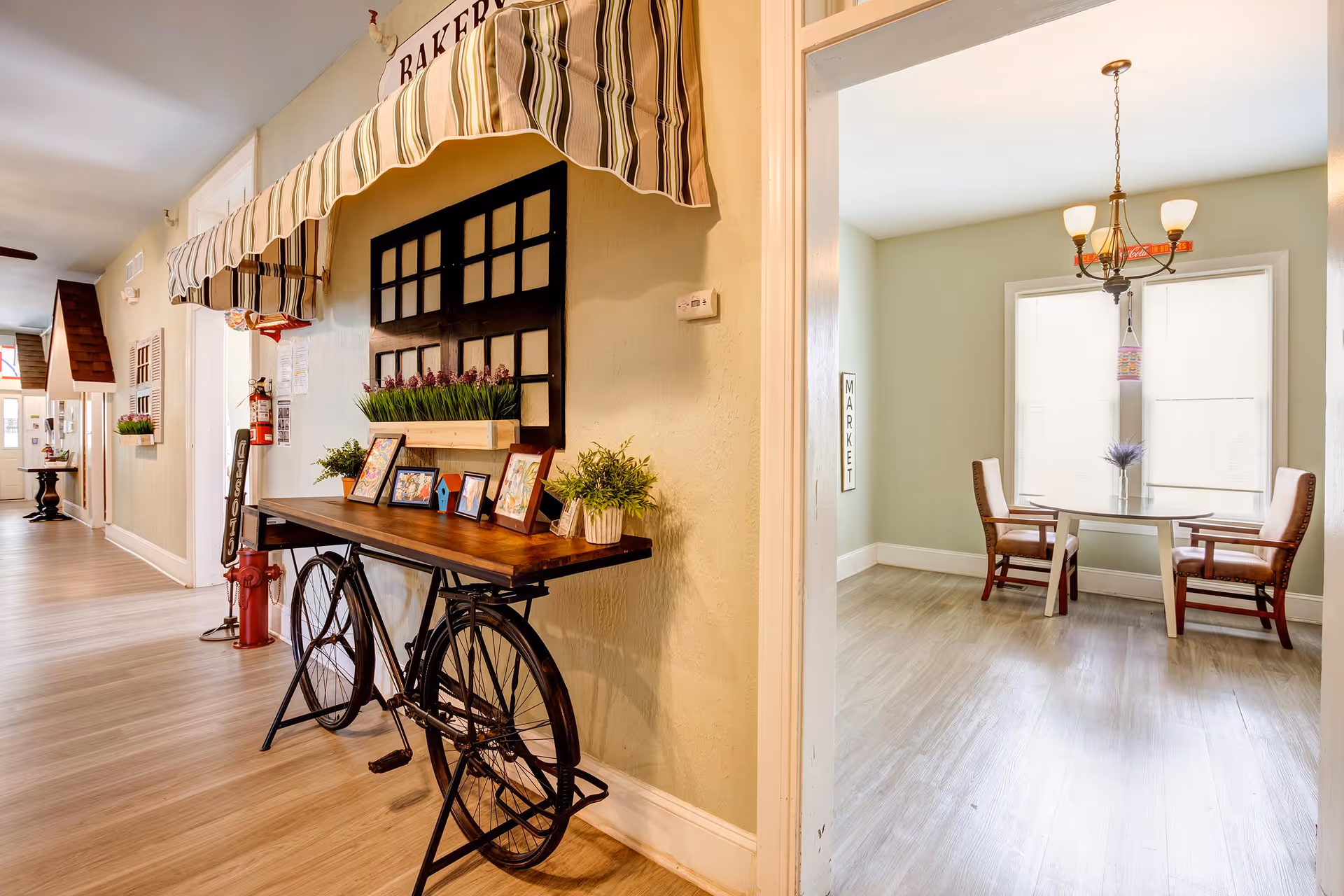 Interior hallway of a senior living facility with a decorative table made from a bicycle frame against the wall, adorned with framed pictures and plants. To the right, there is a small dining area with a round table, two chairs, a chandelier, and large windows with blinds.
