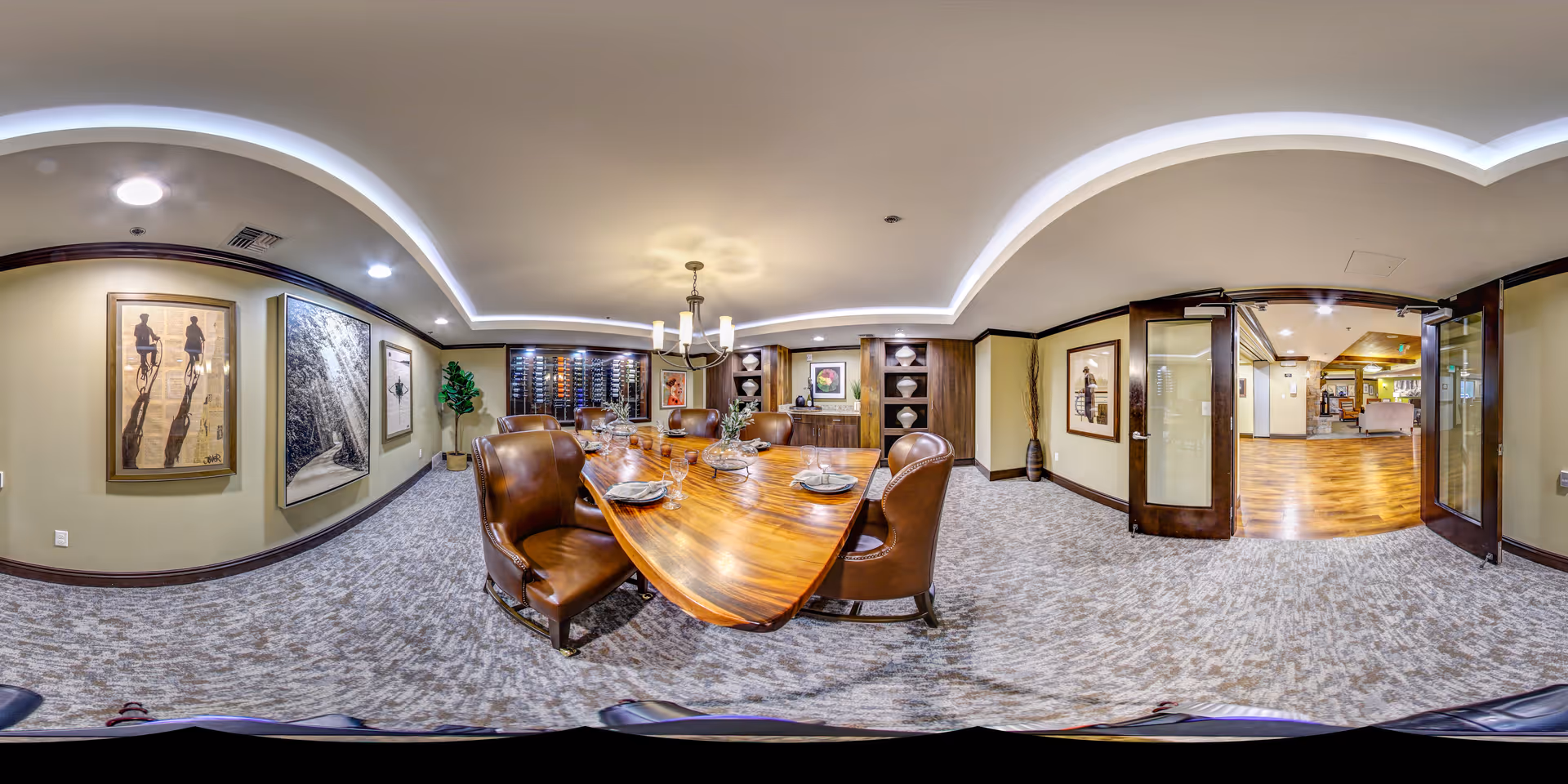 A spacious and well-lit dining room with a large wooden dining table set with plates and glassware. The room features leather chairs, carpeted flooring, framed artwork on the walls, a chandelier, and built-in wooden cabinets with decorative items. There are double glass doors leading to another room with wooden flooring.
