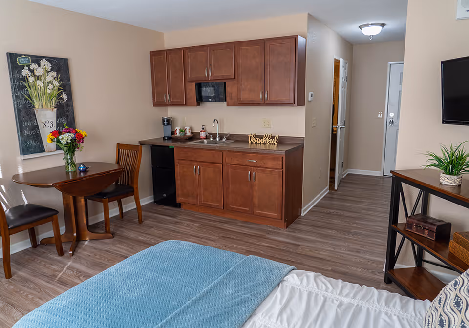 Interior view of a senior living facility room featuring a bed with a blue blanket in the foreground, a small wooden dining table with two chairs and a vase of flowers, a kitchenette with wooden cabinets, a sink, a microwave, and a coffee maker. There is a wall-mounted TV on the right and a small shelf with decorative items. The room has wood flooring and beige walls.