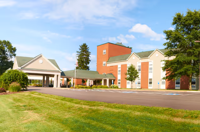 Exterior view of a senior living facility named Suites At Rouse, showing a brick and beige building with green roofs, a covered entrance, a parking lot with handicap spaces, and surrounding green lawn and trees under a blue sky with some clouds.
