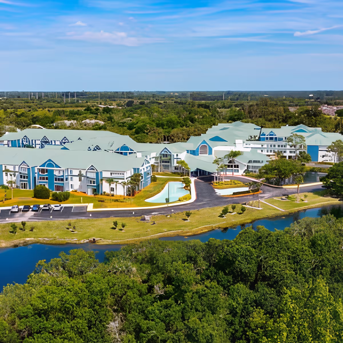 Aerial view of Discovery Village Vero Beach senior living facility surrounded by lush greenery and a small body of water, with multiple white buildings featuring teal roofs under a partly cloudy blue sky.