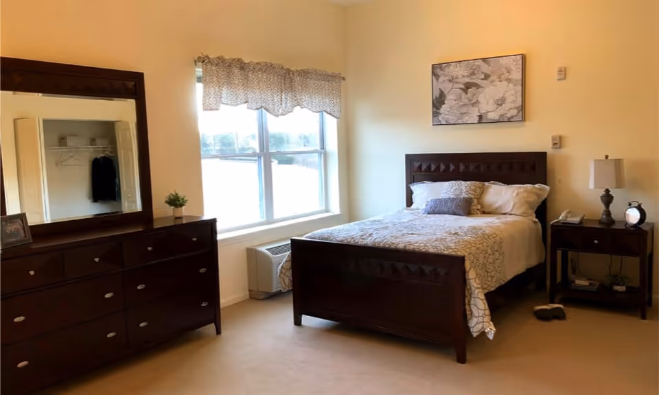 A bedroom with a wooden bed frame and a neatly made bed with white and beige bedding. There is a wooden dresser with a large mirror on the left side and a window with a patterned valance letting in natural light. A nightstand with a lamp, clock, and telephone is on the right side of the bed. The walls are painted a light beige color and there is a framed floral artwork above the bed.