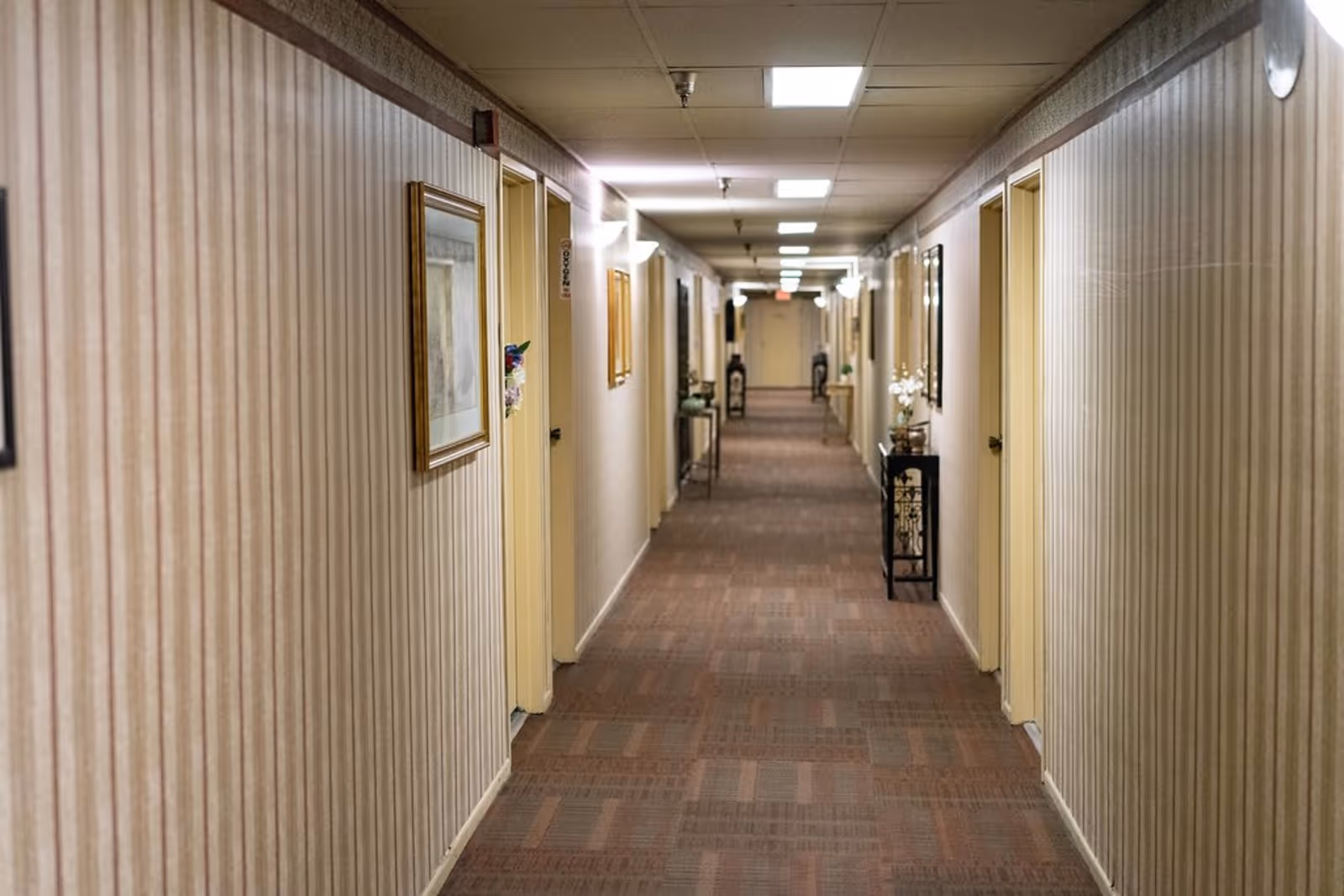 A long, carpeted hallway in a retirement residence with beige striped wallpaper, several closed doors on both sides, framed pictures on the walls, and small tables with decorative items along the corridor. The ceiling has recessed lighting and wall-mounted lamps.