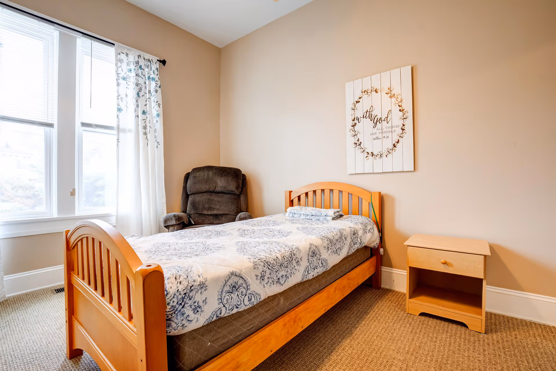 A simple bedroom with a wooden single bed covered with a white and blue patterned bedspread, a folded towel on top, a wooden nightstand with a drawer, a dark brown recliner chair, and a window with white and floral curtains letting in natural light. A decorative wall art with a wreath and text hangs above the bed.