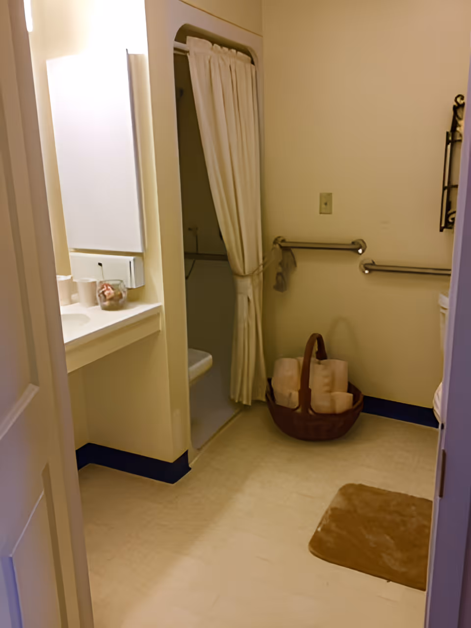 Interior view of a bathroom with a small shower area enclosed by a white curtain, a countertop with a sink and toiletries, grab bars on the wall, a basket with rolled towels on the floor, and a brown bath mat.