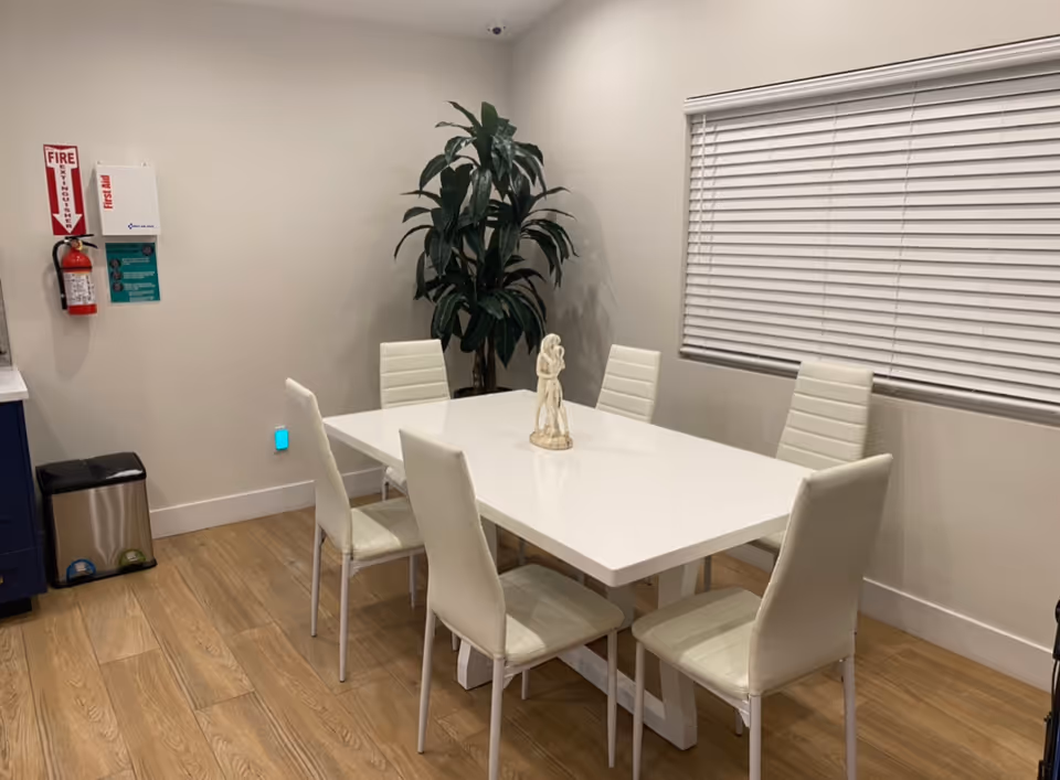 A clean dining area with a white rectangular table surrounded by six white chairs. A small decorative statue is placed in the center of the table. There is a large green plant in the corner near the wall, a window with closed white blinds, a fire extinguisher mounted on the wall, and a stainless steel trash bin on the wooden floor.