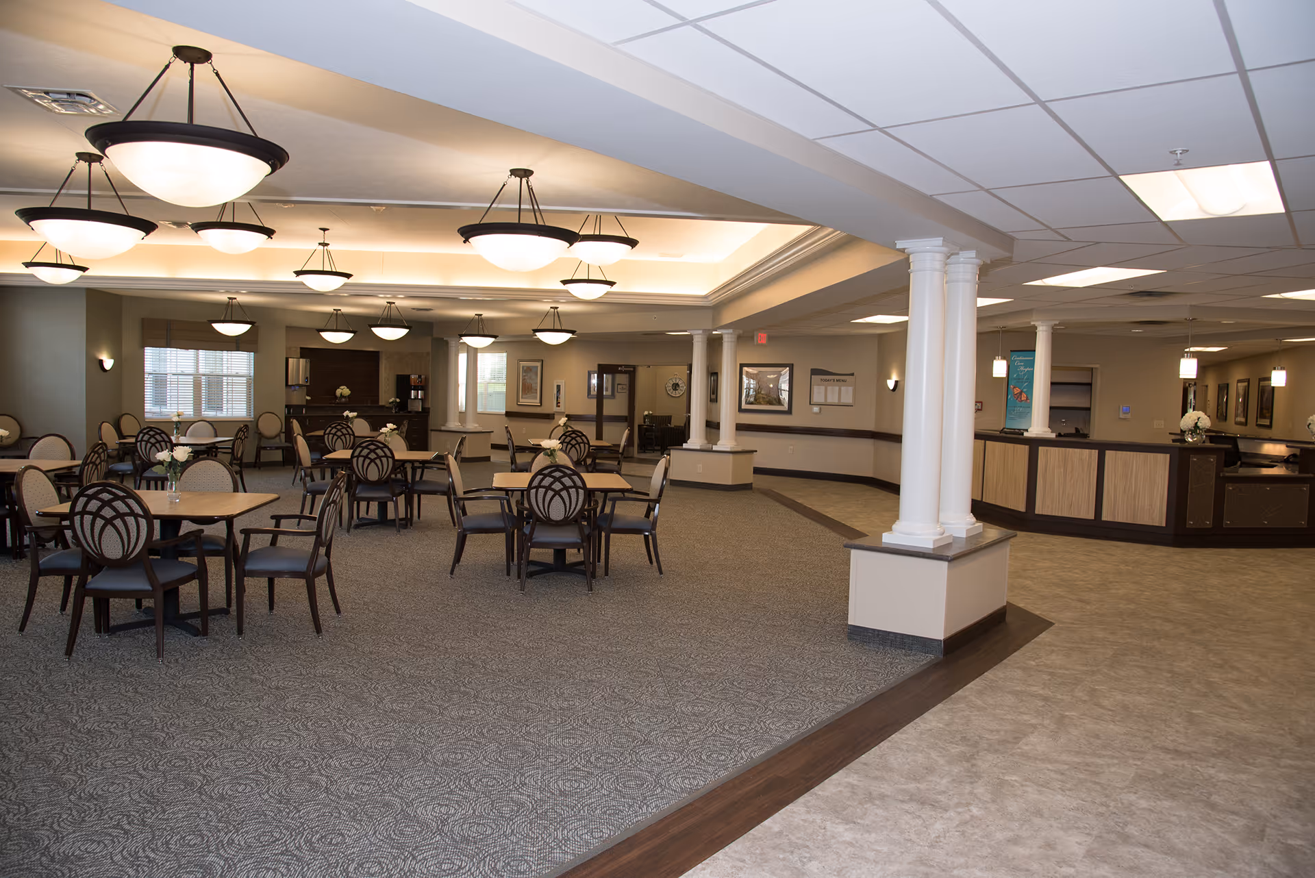 A spacious dining area in a senior living facility with multiple round tables and chairs arranged neatly. The room features carpeted flooring, several large ceiling lights, white columns, and a reception or service counter area to the right. The walls are decorated with framed pictures and windows with blinds.
