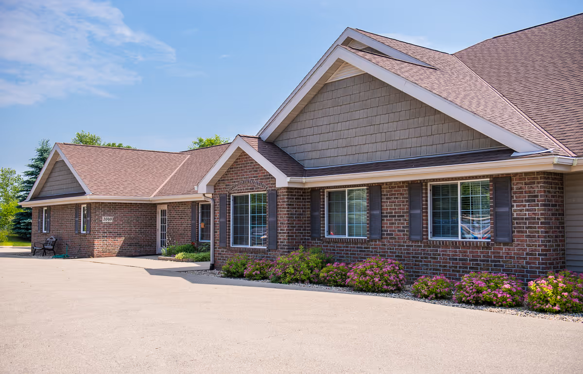 Exterior view of a single-story brick building with multiple windows, brown roof shingles, and flower beds along the front. The sky is clear with some clouds.