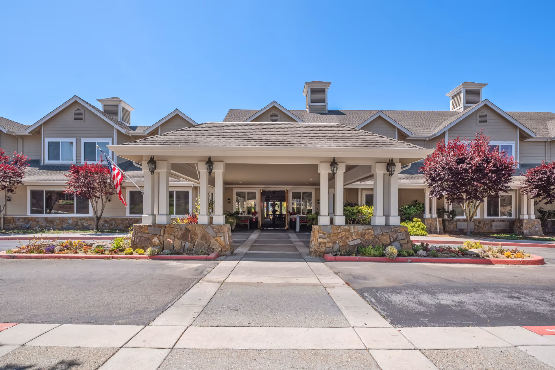 Front entrance of a senior living facility with a covered driveway, stone pillars, and beige siding. There are red-leaved trees and landscaped flower beds on either side of the entrance, and an American flag is displayed on the left side. The sky is clear and blue.
