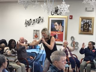 A group of elderly people seated in a room, some raising their hands and smiling. A woman is standing and holding a microphone, engaging with the group. The room is decorated with musical notes, snowflakes, and a framed poster of Elvis Presley on the wall.