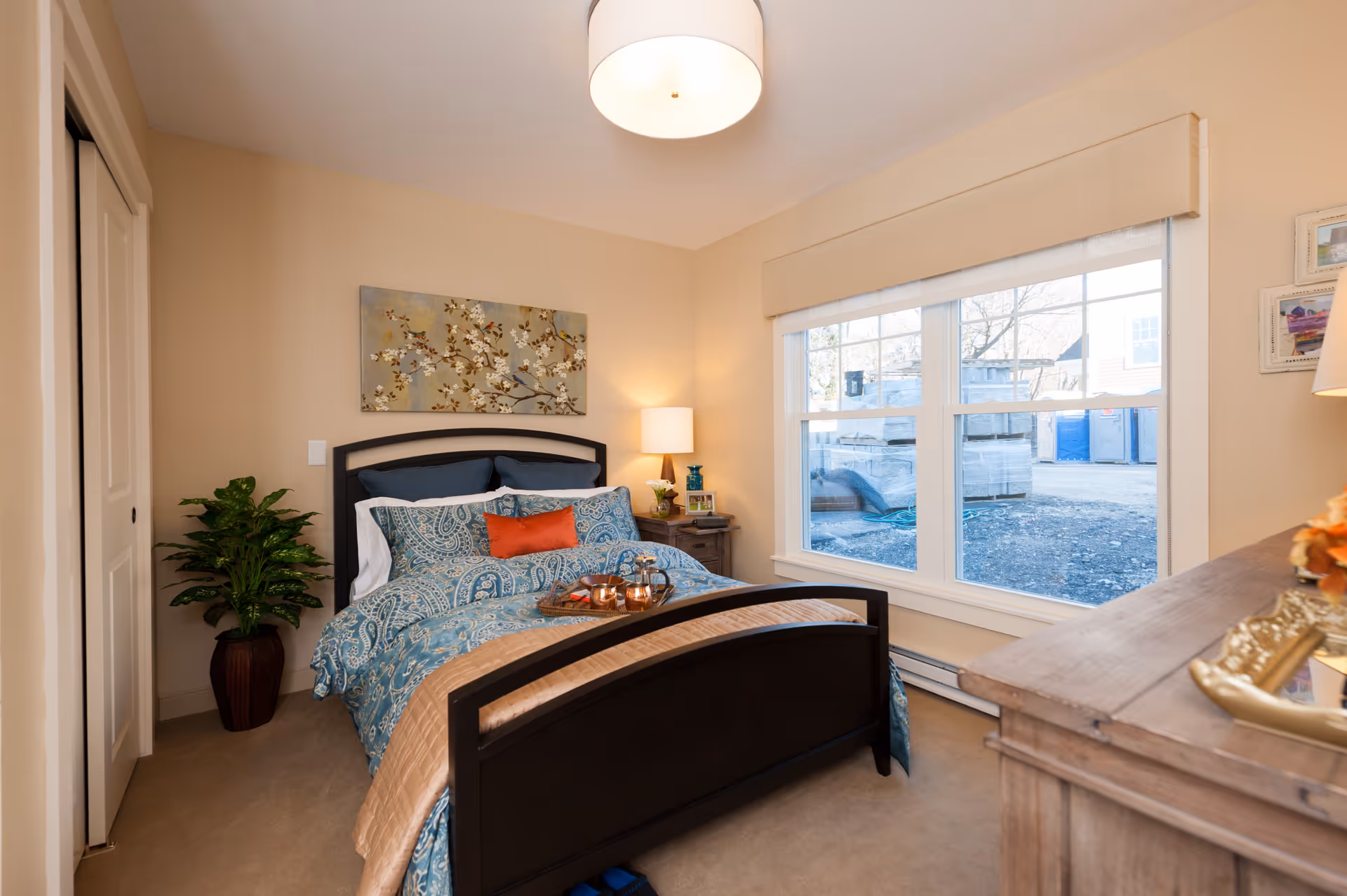 A cozy bedroom with a dark wooden bed frame, blue patterned bedding, and an orange accent pillow. There is a nightstand with a lamp and decorative items next to the bed, a large window with a beige valance letting in natural light, a potted plant in the corner, and a dresser with decorative items on the right side. The walls are painted beige and there is a floral artwork hanging above the bed.
