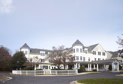 Exterior view of a large, multi-story senior living facility with white siding, multiple peaked roofs, and a covered entrance. The building is surrounded by trees and a white fence, with a paved driveway and lawn in the foreground under a clear sky.