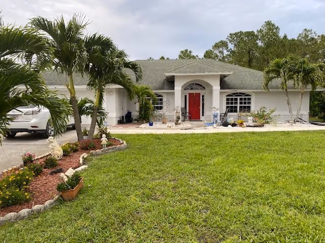 Front exterior view of a single-story house with a green lawn, palm trees, and a red front door. There is a driveway with a white car parked on the left side and various plants and garden decorations near the entrance.