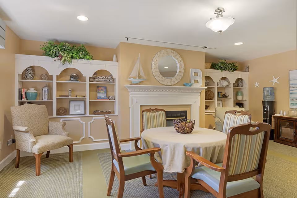 A cozy living room area with a round table covered with a beige tablecloth surrounded by four wooden chairs with striped cushions. Behind the table is a white fireplace with a round decorative mirror above it and a model sailboat on the mantel. On either side of the fireplace are built-in shelves decorated with various ornaments, books, and plants. To the left is a beige upholstered armchair, and to the right is a water cooler and a wooden cabinet. The walls are painted a warm beige color, and the room is well-lit with ceiling lights.