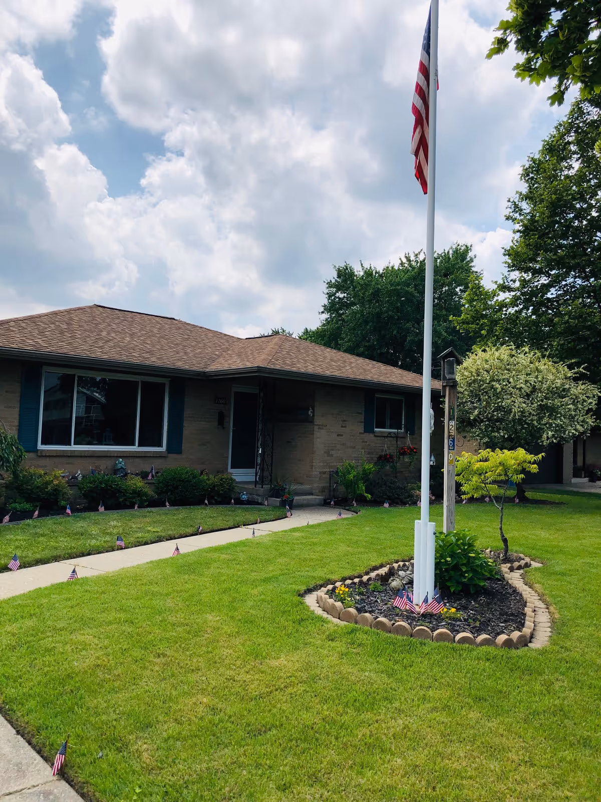 Single-story brick building with a brown shingled roof, surrounded by a well-maintained lawn with small American flags lining the walkway and a flagpole with an American flag in the front yard. Trees and bushes are visible around the property under a partly cloudy sky.