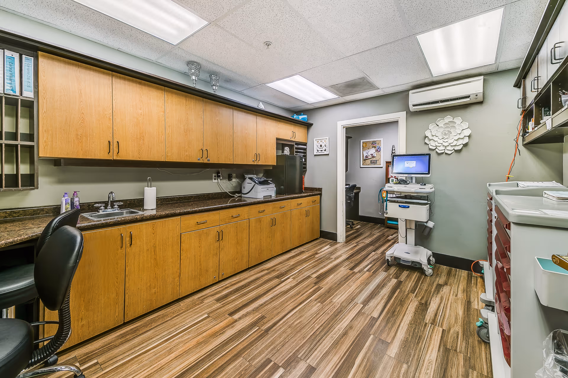 A clean and organized medical or clinical workspace with wooden cabinets along the left wall, a countertop with a sink, and various office and medical equipment. The room has wood-patterned flooring, overhead fluorescent lighting, and a computer workstation on a mobile cart near the doorway. The walls are painted light gray with a decorative white flower wall hanging.