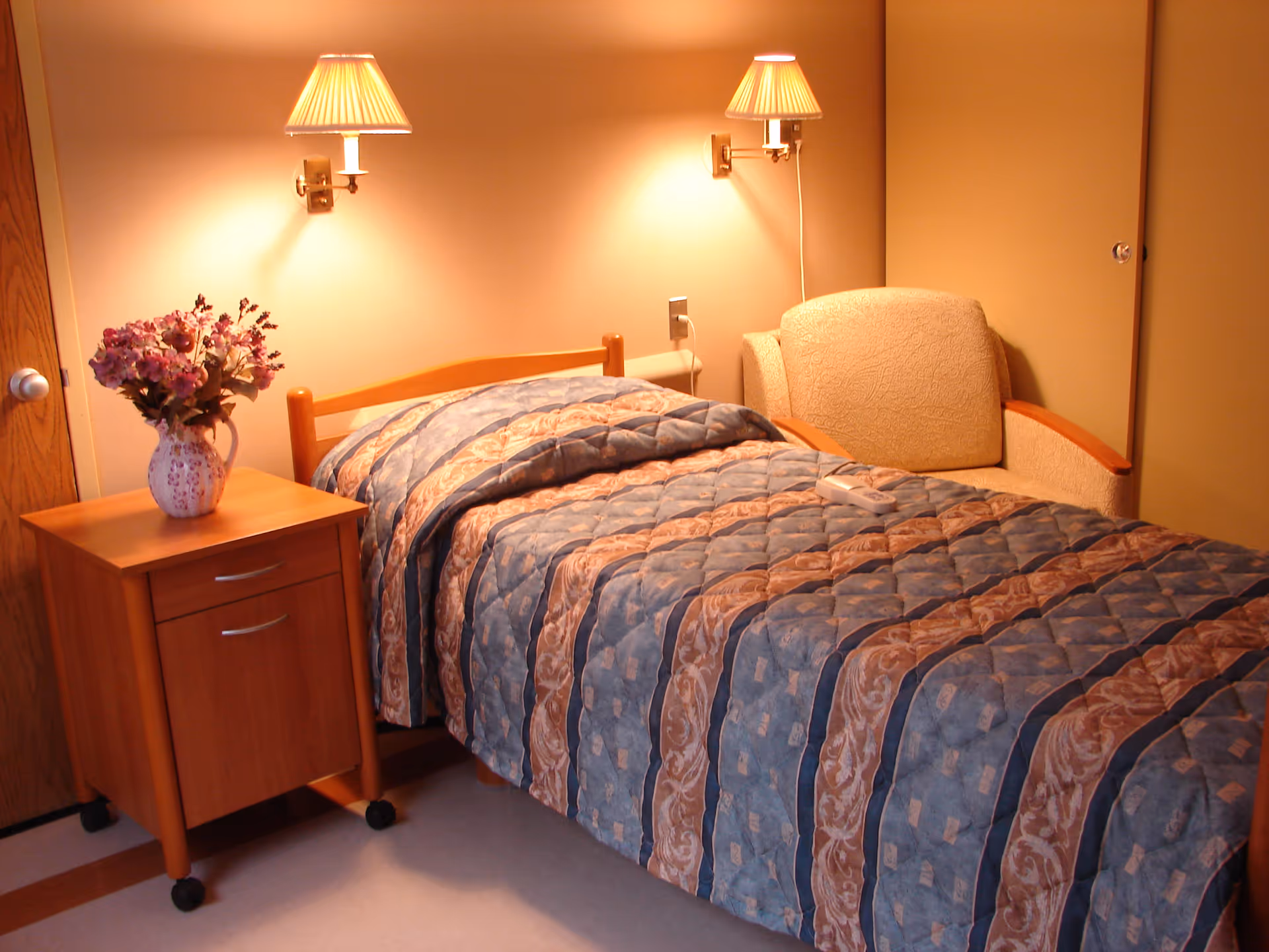 A neatly made single bed with a patterned quilt in a room. Next to the bed is a wooden nightstand with a vase of purple flowers. Two wall-mounted lamps with beige lampshades provide warm lighting. There is a beige upholstered armchair beside the bed and a closed wooden door in the background.