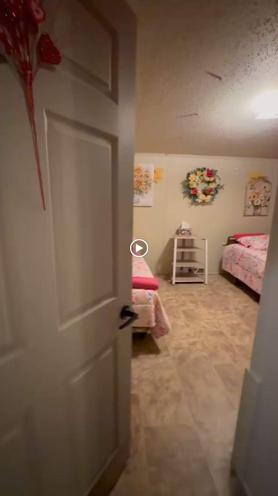 View through a partially open door into a bedroom with two beds covered in floral bedspreads. A small white nightstand with a tissue box is positioned between the beds. The walls are decorated with floral artwork and a flower wreath. The floor has a wood-like finish.