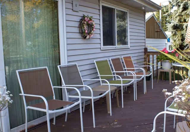 Outdoor patio area with six metal-framed chairs with different colored seats arranged in a row against the exterior wall of a building. The wall has horizontal siding, a window, and a decorative wreath. There are potted plants on the patio and greenery visible in the background.