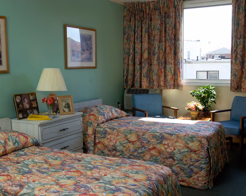 A bedroom with two twin beds covered in floral patterned bedspreads. Between the beds is a white nightstand with a lamp, framed photos, and a small vase with flowers. The room has green walls with framed artwork and a window with floral curtains letting in natural light. Two blue armchairs and a small table with a plant and flowers are near the window.