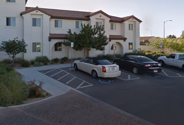 Exterior view of a two-story assisted living facility building with white walls and a red-tiled roof. There are several windows and arched doorways. In front of the building is a parking lot with three cars parked, including two in handicapped spaces. There are small trees and shrubs around the building and parking area.