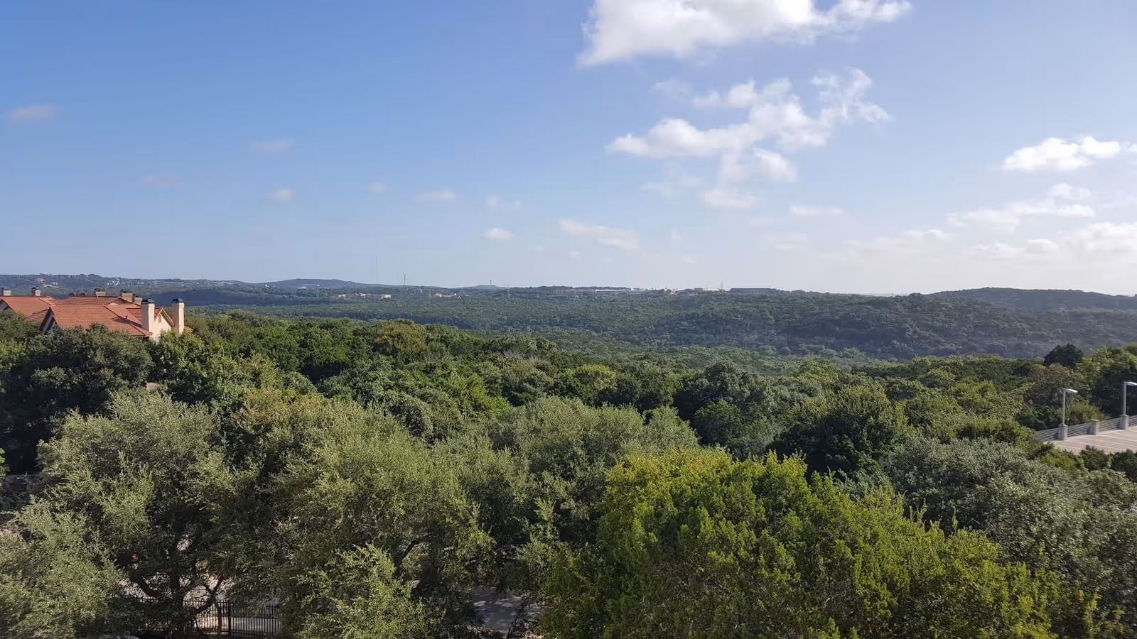 A scenic view of a lush green forest with a clear blue sky and scattered clouds. In the distance, there are hills and a few rooftops of buildings with red tiles visible on the left side.