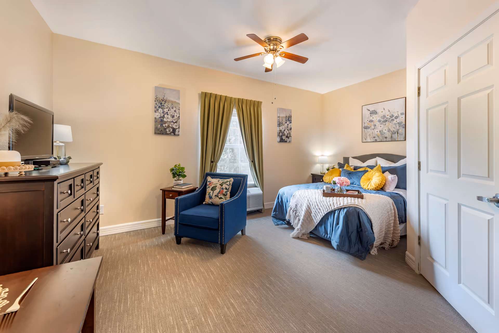 A cozy bedroom with a bed dressed in blue and white bedding, accented with yellow pillows. There is a blue armchair with a decorative pillow, a wooden dresser with a TV on top, and a small side table with a plant. The room has beige walls, a window with green curtains, and floral artwork on the walls. A ceiling fan with lights is mounted on the ceiling.