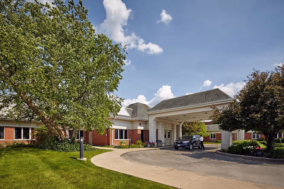 Front exterior view of Oak Brook Healthcare Centre showing a covered entrance with a car parked underneath and a person standing beside it. The building has red brick walls and white trim, surrounded by green trees and a well-maintained lawn under a partly cloudy blue sky.