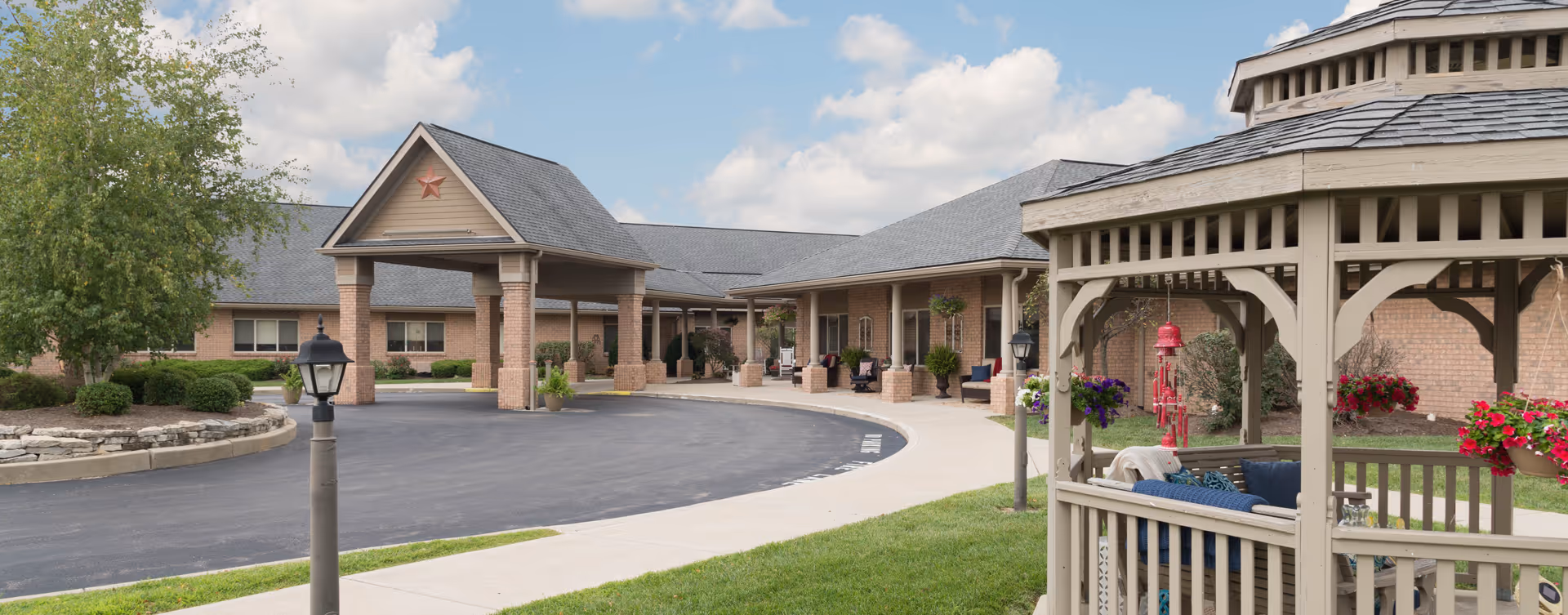 Exterior view of Bickford of Middletown senior living facility showing a circular driveway, covered entrance with columns, a gazebo with seating, hanging flower baskets, and well-maintained landscaping under a partly cloudy sky.