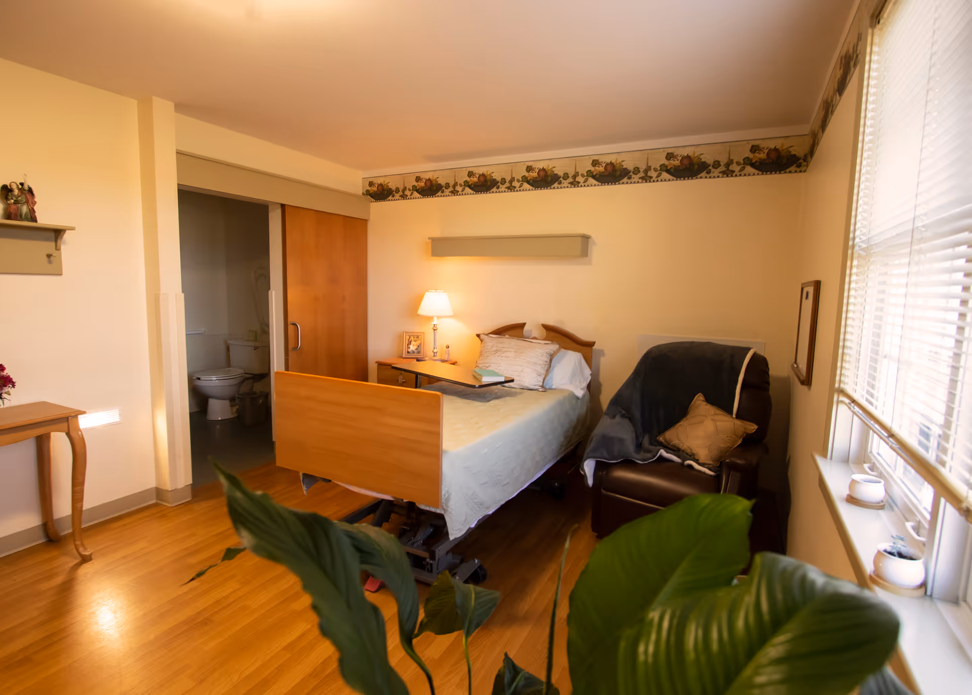 A cozy nursing home bedroom with a hospital bed, a side table with a lamp and picture frame, a comfortable armchair with a blanket and pillow, a window with blinds, and a partially visible bathroom with a toilet. The room has wooden flooring and light-colored walls with a decorative border near the ceiling.