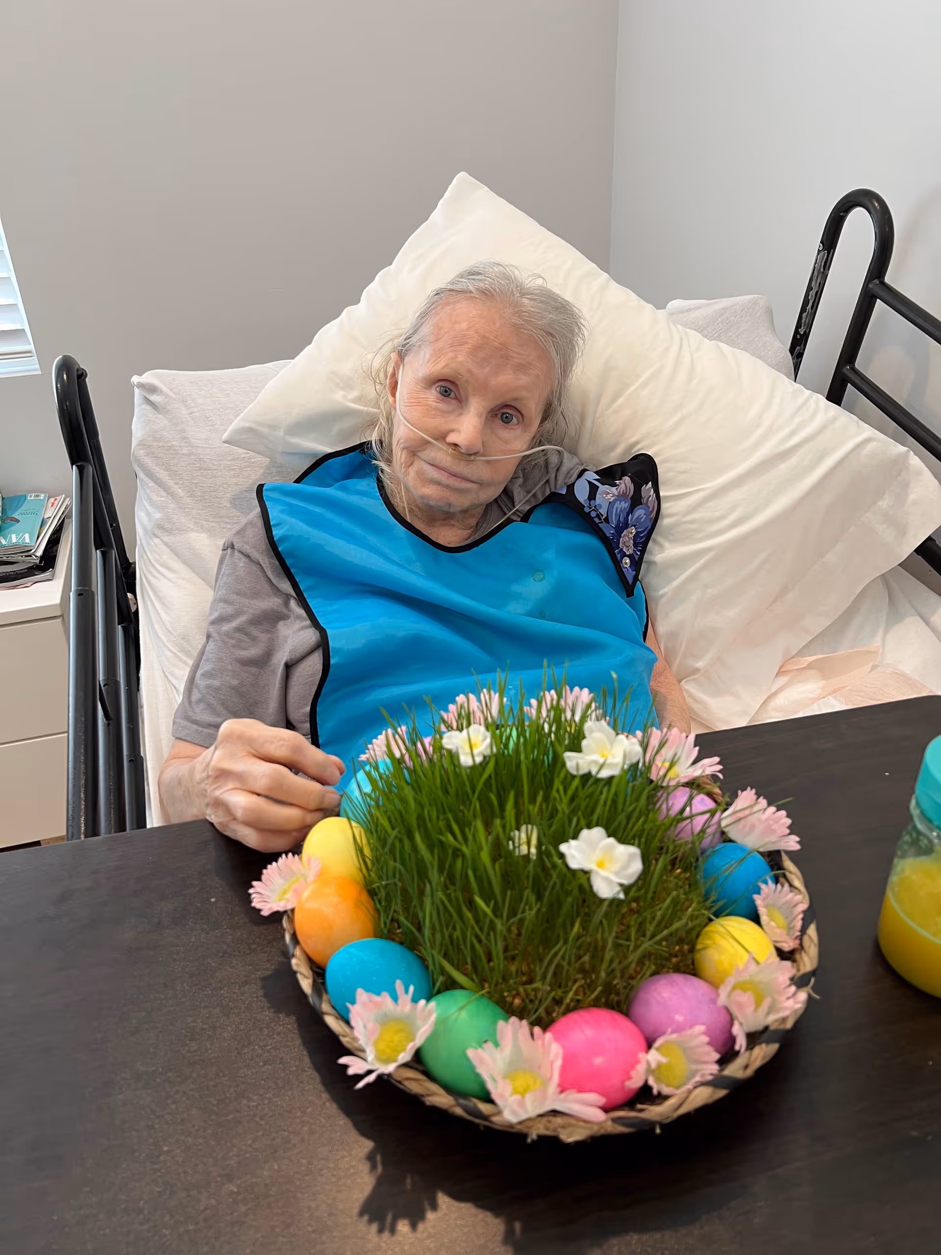 An elderly woman lying in a hospital-style bed with a nasal oxygen tube, wearing a blue bib, and sitting up against white pillows. In front of her on a table is a decorative basket with green grass, white flowers, colorful Easter eggs, and pink flowers. A jar with orange juice is also visible on the table.