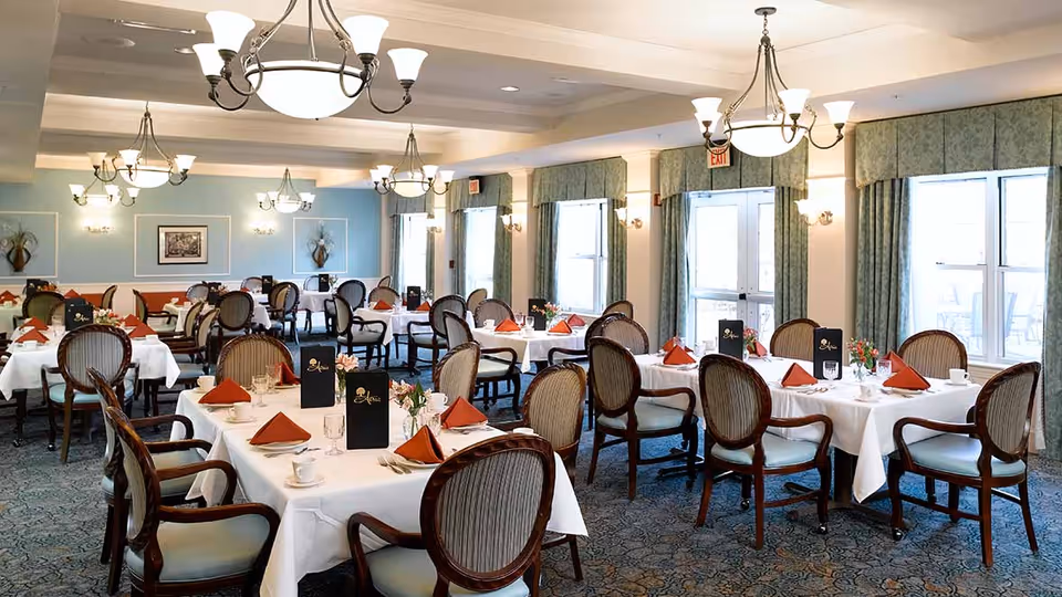 A spacious, well-lit dining room with multiple round and rectangular tables covered in white tablecloths. Each table is set with orange folded napkins, glassware, cups, and small floral centerpieces. The room features elegant wooden chairs with cushioned seats and backs, blue patterned carpet, light blue walls with framed artwork, and large windows with green curtains allowing natural light to enter. Several chandeliers and wall sconces provide additional lighting.